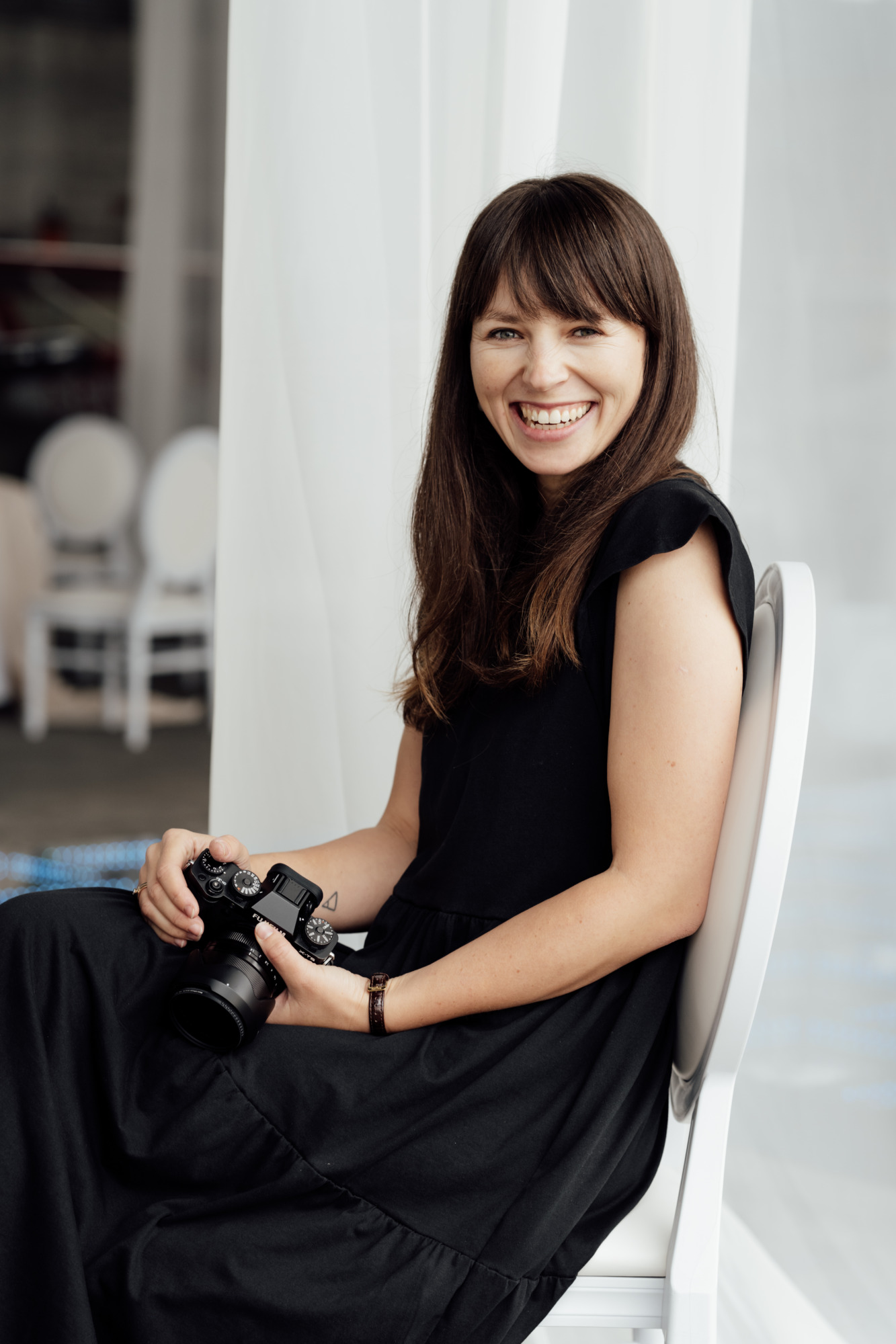 Professionelle Hochzeitsfotografin, die lächelnd mit Kamera in weichem Tageslicht sitzt.
