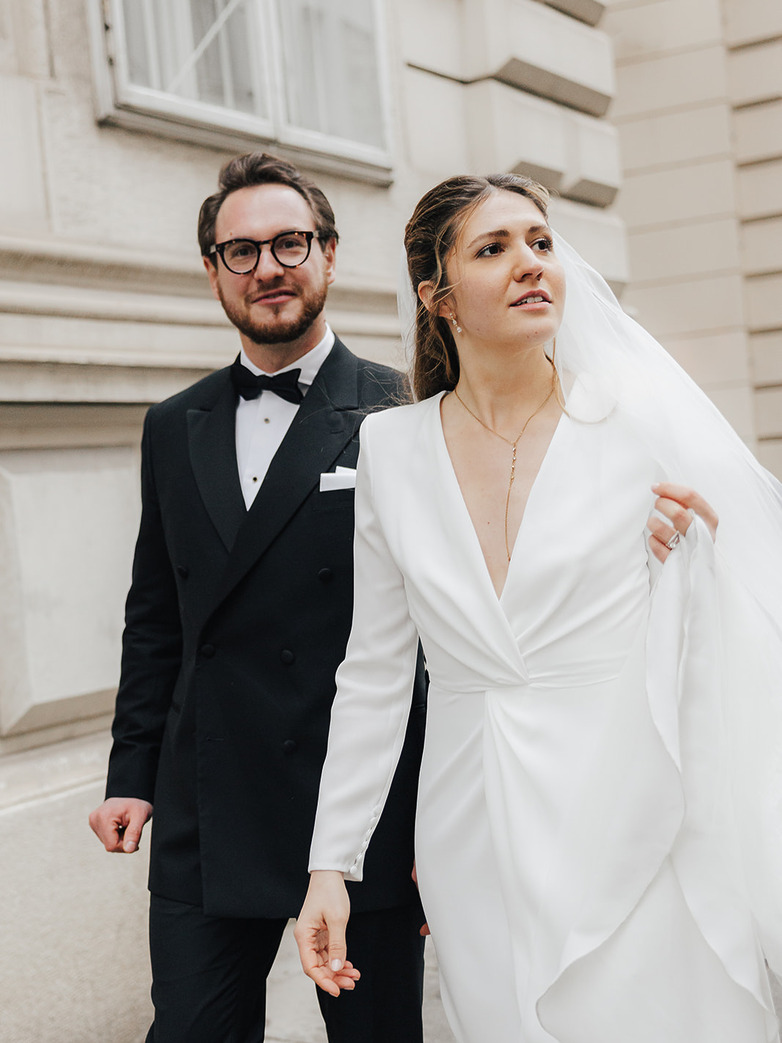 Bride adjusting her veil while walking beside the groom during an elegant couple shoot in Vienna, both wearing modern black tie wedding attire.