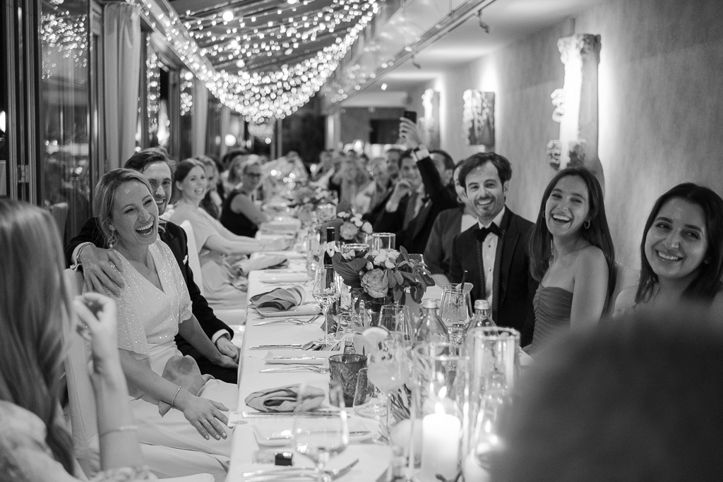Black and white photo of laughing guests at a long table under string lights at the Settimo Cielo rooftop in Vienna