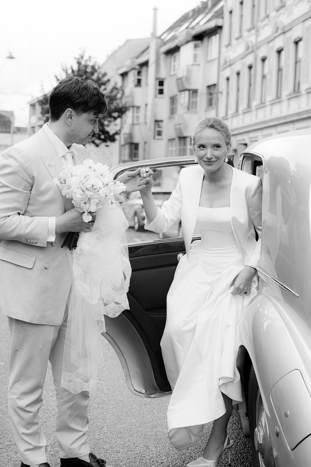 Black and white photo of a bride stepping out of a vintage car with the groom on a Vienna street